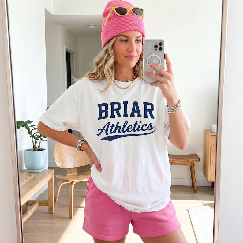 May include: A white short-sleeved t-shirt with the text BRIAR Athletics printed in dark blue, paired with pink shorts and a matching pink beanie. The t-shirt features a block letter font for BRIAR and a cursive script font for Athletics.