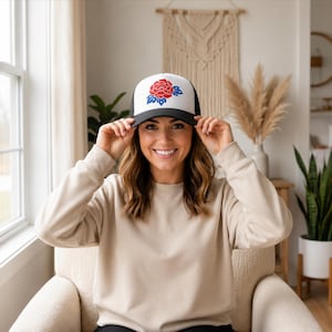 May include: A trucker cap featuring a white front panel with a red and blue floral graphic design. The cap has a black peak and black mesh side and back panels. The design depicts a stylised flower with red petals and blue leaves.