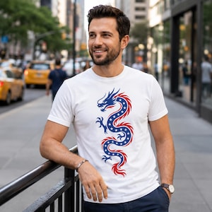 May include: A white short-sleeved t-shirt featuring a central graphic of a serpentine dragon. The dragon is coloured in blue with white stars and outlined in red, creating a stylised design that runs vertically down the centre of the chest area.