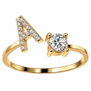 May include: A gold open ring featuring a letter A encrusted with small clear stones on one end and a single round clear stone set in a four-prong mount on the other end. The band is smooth and polished, creating a continuous circular shape.