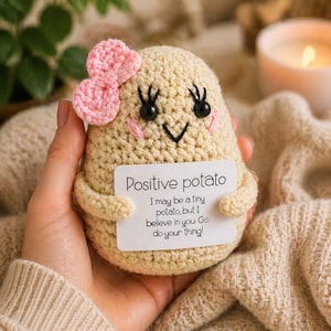 Può includere: Un peluche a forma di patata all'uncinetto beige con un fiocco di lana rosa, occhi a perla neri e ciglia ricamate. Tiene in mano un cartoncino bianco con il testo: Positive potato. I may be a tiny potato, but I believe in you Go do your thing!