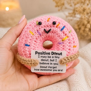 Könnte beinhalten: Ein gehäkelter rosa Donut mit kleinen schwarzen Perlenaugen und einem gestickten schwarzen Mund. Der Donut ist mit bunten Garnstreuseln verziert und hält eine weiße rechteckige Karte mit dem Text: Positive Donut, I may be a tiny donut, but I believe in you. Donut forget how awesome you are!