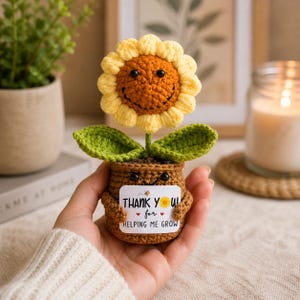 Op de afbeelding: Een gehaakte zonnebloem met een bruin hart, gele bloemblaadjes en twee groene bladeren, geplant in een bruine gehaakte pot. De pot heeft een klein lachend gezichtje en houdt een wit bordje vast met de tekst THANK YOU for HELPING ME GROW.