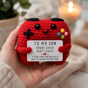 Puede incluir: Un peluche de mando de juego tejido a ganchillo en rojo con detalles de botones negros y pequeños acentos circulares de colores. Sostiene una tarjeta rectangular blanca que dice: TO MY SON YOU'RE LOVED NEXT LEVEL! A TINY GAME CONTROLLER IS HERE TO ALWAYS REMIND YOU!