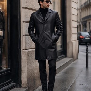 May include: A black leather overcoat featuring a notched lapel, three front buttons, and side pockets. The coat is worn over a black turtleneck sweater and black trousers, paired with black leather dress shoes and dark-tinted sunglasses.
