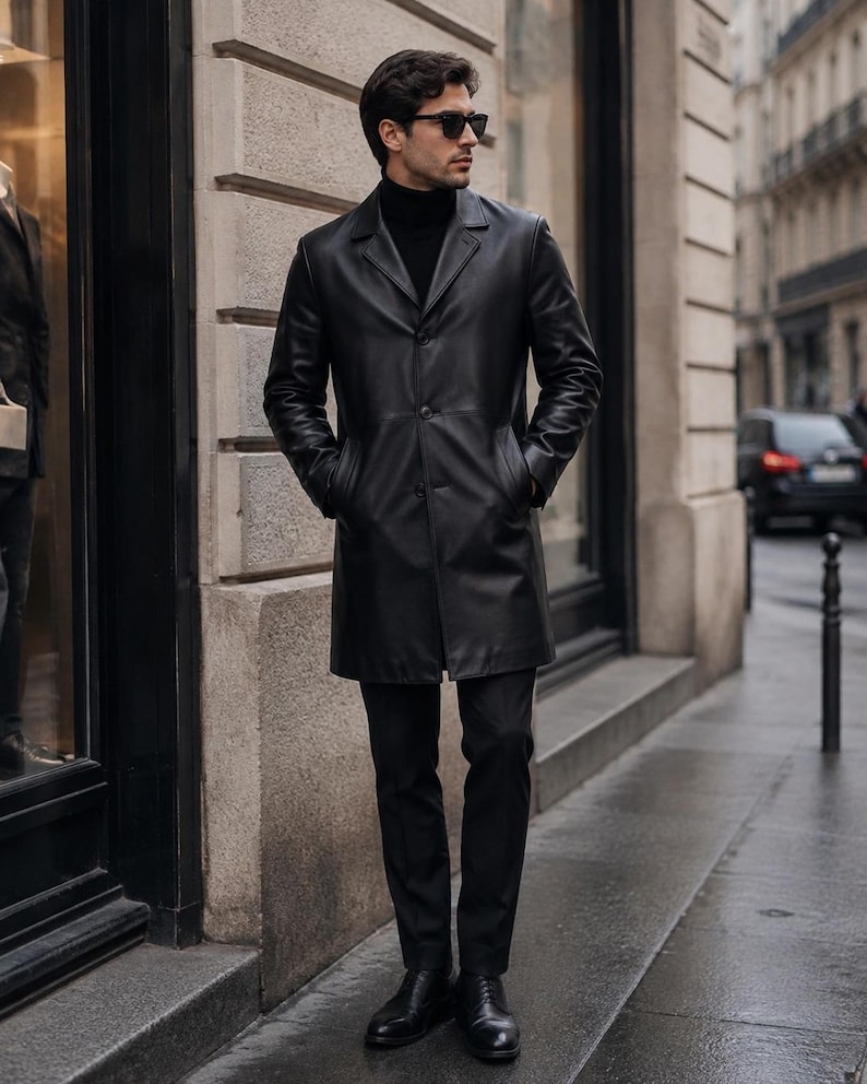 May include: A black leather overcoat featuring a notched lapel, three front buttons, and side pockets. The coat is worn over a black turtleneck sweater and black trousers, paired with black leather dress shoes and dark-tinted sunglasses.