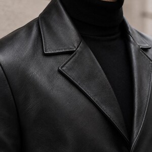 May include: A black leather blazer with a notched lapel collar and a single visible button closure, worn over a black ribbed turtleneck sweater. The jacket features a smooth, matte-textured finish with visible stitching along the lapel edges.
