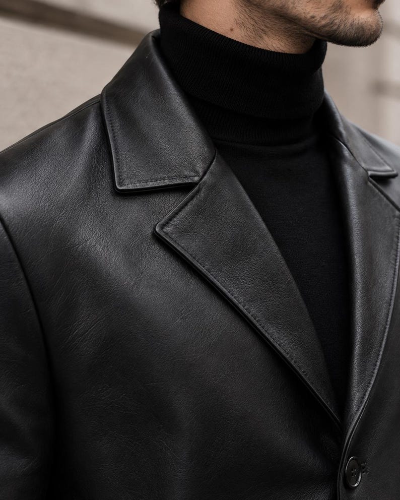 May include: A black leather blazer with a notched lapel collar and a single visible button closure, worn over a black ribbed turtleneck sweater. The jacket features a smooth, matte-textured finish with visible stitching along the lapel edges.