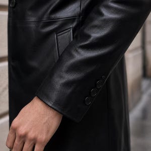 May include: A black leather coat featuring a smooth texture, a single button closure on the front, and a sleeve cuff detailed with four small, matching black buttons arranged in a vertical row along the outer edge of the wrist area.