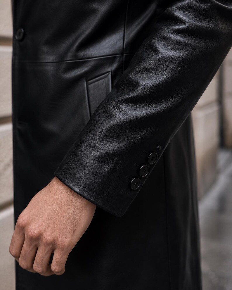 May include: A black leather coat featuring a smooth texture, a single button closure on the front, and a sleeve cuff detailed with four small, matching black buttons arranged in a vertical row along the outer edge of the wrist area.
