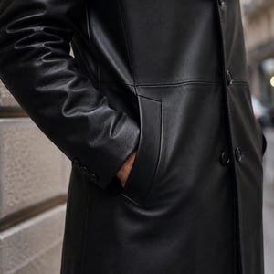 May include: A long black coat made of smooth material featuring a front button closure, long sleeves with three small buttons at each cuff, and deep side pockets. The garment has a structured collar and a clean, minimalist design throughout.