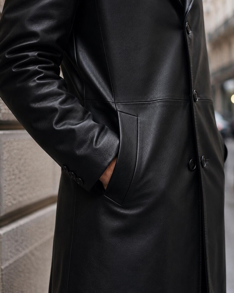 May include: A long black coat made of smooth material featuring a front button closure, long sleeves with three small buttons at each cuff, and deep side pockets. The garment has a structured collar and a clean, minimalist design throughout.