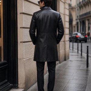 May include: A black leather trench coat featuring a structured collar, long sleeves, and a smooth finish. The coat reaches mid-thigh and is worn over black trousers and black shoes. The garment is shown from the back, displaying a central vent.