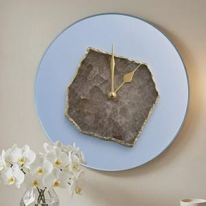 May include: A round wall clock with a light blue face and a central, irregular, dark brown stone slab featuring a gold-toned rim. The clock has two gold-toned hands positioned at the center of the stone, mounted on a wall above a white table.