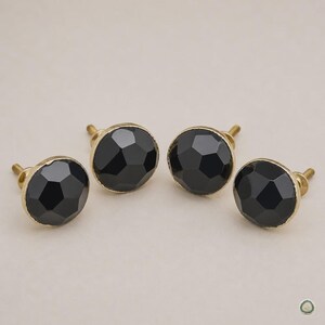 May include: Four round cabinet knobs featuring faceted black surfaces and gold-toned metal bases. Each knob has a circular, multi-faceted top that reflects light, attached to a short, threaded metal post for installation on furniture or doors.