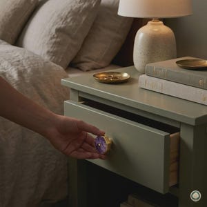 May include: A sage green wooden nightstand featuring a single drawer with a hexagonal purple amethyst stone knob with a gold-toned metal edge. The nightstand is topped with a speckled ceramic lamp and two stacked books with a small gold tray.