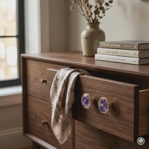 May include: A dark wood dresser with a partially open drawer featuring two hexagonal purple amethyst knobs with gold-toned edges. A light-colored patterned silk scarf hangs over the drawer front. Three stacked books and a ceramic vase sit on top.