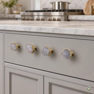 Op de afbeelding: Vier kastknoppen met onregelmatige, doorschijnende grijze steenschijven met getextureerde, metallic goudkleurige randen. Elke knop is gemonteerd op een ronde goudkleurige basis en bevestigd aan een gladde, matgrijze keukenladefront.