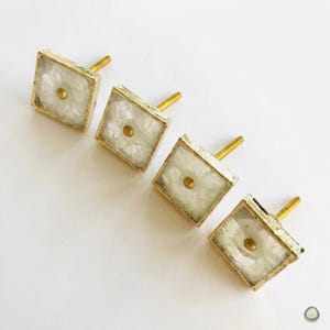 May include: Four square cabinet knobs featuring a translucent white, textured inlay encased in a gold-toned metal frame. Each knob has a centered gold-toned circular stud and a gold-toned mounting post extending from the back for installation.