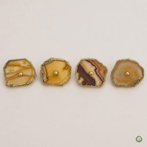 May include: Four cabinet knobs featuring irregular, polished agate slices in shades of tan, cream, and deep purple. Each stone has a textured, metallic gold-toned edge and a small, centered gold-toned metal knob for attachment to furniture.