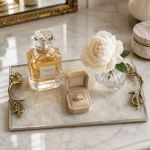 May include: A rectangular white stone tray with gold-toned ornate handles holding a glass bottle of Guerlain perfume, a beige velvet ring box containing a gold ring with a round stone, and a small glass vase with a single white peony flower.