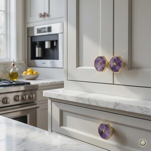 May include: Three octagonal cabinet knobs featuring polished purple amethyst stone centers encased in gold-toned metal frames. These decorative hardware pieces are mounted on light gray kitchen cabinetry with white marble countertops in the background.