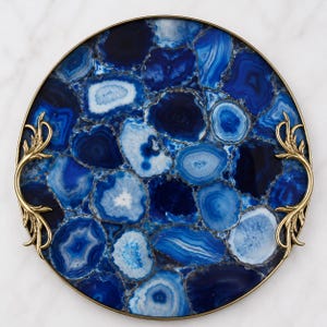 May include: A circular serving tray featuring a mosaic of blue and white agate slices. The tray is encased in a thin gold-toned metal rim and includes two ornate, curved gold-toned metal handles on opposite sides, showcasing intricate leaf-like detailing.