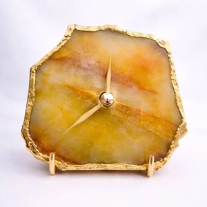 Puede incluir: Un reloj de mesa con una losa irregular y translúcida de piedra ágata en tonos amarillos y ámbar. Los bordes de la piedra tienen un acabado con un borde dorado metálico. Dos manecillas doradas delgadas están montadas en el centro.