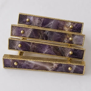 Puede incluir: Un tirador de mueble rectangular con una incrustación de piedra amatista morada pulida, engastada en un marco de metal dorado texturizado. El tirador está asegurado por dos pequeños pernos metálicos redondos dorados en cada extremo.