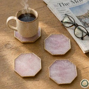 Könnte beinhalten: Ein Set aus drei achteckigen Untersetzern aus Rosenquarz mit goldfarbenen Metallrändern, begleitet von einer gesprenkelten beigen Keramiktasse mit schwarzem Kaffee und einer schwarz umrandeten Brille, die auf einer Zeitung liegt.