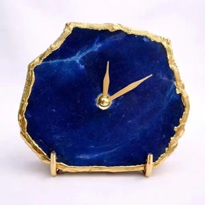 May include: A desk clock featuring a polished, deep blue agate slice as the face, framed by a textured gold-colored metallic edge. The clock has two slender gold-colored hands at the center and rests on a small, gold-colored metal stand.