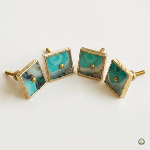 May include: Four square cabinet knobs featuring natural teal and white agate slices encased in gold-toned metal frames. Each knob has a central gold-toned stud and a threaded metal bolt extending from the back for installation on furniture.