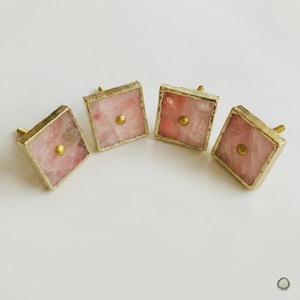 May include: Four square cabinet knobs featuring translucent pink rose quartz stone centers encased in textured gold-toned metal frames. Each knob has a small, centered gold-toned circular stud detail on the front surface of the stone.