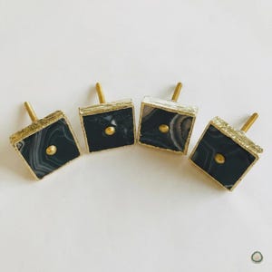 Op de afbeelding: Vier vierkante kastknoppen met zwart-wit gestreepte agaatstenen oppervlakken. Elke knop heeft een metallic goudkleurige rand en een kleine goudkleurige centrale knop. De knoppen zijn in een horizontale rij op een witte achtergrond geplaatst.