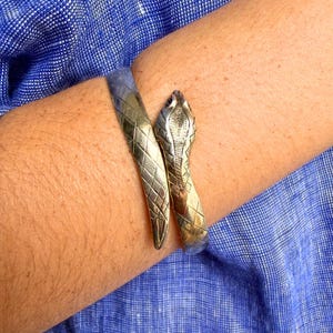 May include: A metallic gold-toned snake-shaped cuff bracelet featuring a textured diamond-patterned body and a detailed cobra head design. The bracelet wraps around the wrist, showcasing engraved scales and a polished finish on the metal surface.