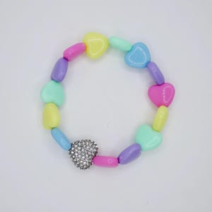 May include: A stretch bracelet composed of smooth, heart-shaped beads in pastel shades of pink, yellow, purple, blue, and mint green. One heart-shaped bead is covered in small, clear, sparkling rhinestones, creating a focal point on the band.