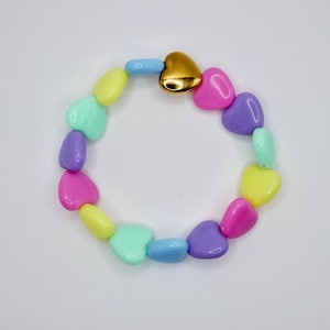 Può includere: Un braccialetto circolare composto da perline lisce a forma di cuore in vari colori pastello, tra cui rosa, giallo, viola e azzurro. Una singola perlina metallica dorata a forma di cuore è posizionata nella parte superiore del braccialetto, in contrasto con la finitura opaca delle altre perline colorate.