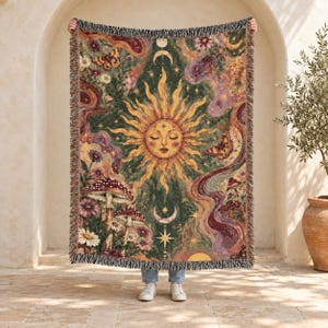 Peut inclure: Un plaid tissé présentant un visage de soleil central aux yeux fermés, entouré de motifs célestes incluant lunes, étoiles, papillons et champignons. Le design intègre une riche palette de couleurs vert, or et violet.