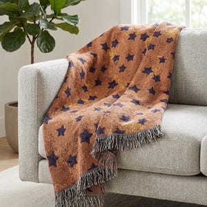 May include: A woven throw blanket featuring a repeating pattern of dark navy blue stars against a textured, mottled rust-orange and gold background. The blanket has a fringed edge with alternating light and dark threads along the bottom border.