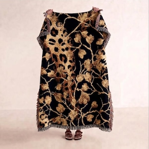 May include: A rectangular woven throw blanket featuring a black background with a gold and brown leopard print pattern intertwined with golden tree branches and leaves. The blanket has a fringed edge along the sides and bottom hem.