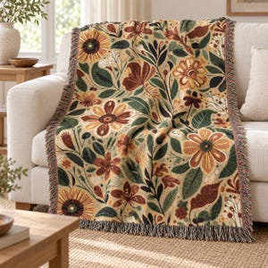 Op de afbeelding: Een geweven plaid met een herhalend bloemenpatroon met grote zonnebloemen, kleinere bloesems en bladeren in tinten mosterdgeel, diep roestoranje, donker bosgroen en crème op een lichtbruine achtergrond.