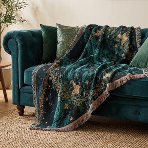 May include: A woven throw blanket featuring a dark teal and black background with intricate gold-colored botanical and butterfly patterns. The rectangular blanket has a fringed edge and is draped over a deep teal velvet sofa with matching pillows.