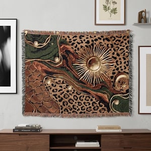 May include: A rectangular woven tapestry featuring a collage of patterns including leopard print, cracked earth textures, and deep green swirls. A prominent gold sunburst emblem sits centrally, accompanied by a crescent moon and small star motifs.