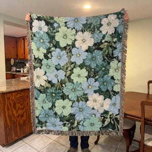 May include: A woven throw blanket featuring a dense floral pattern in shades of light blue, sage green, and white against a black background. The rectangular blanket has a textured finish and is finished with decorative fringe along the edges.