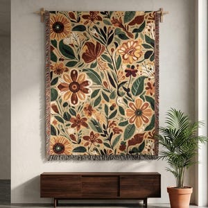 Puede incluir: Una manta tejida rectangular con un denso patrón botánico de flores y hojas estilizadas en tonos óxido, amarillo mostaza, verde oscuro y crema sobre un fondo beige, terminada con un borde de flecos en la parte inferior.