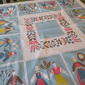 Könnte beinhalten: Vintage Tischdecke mit hellblauem und weißem Design, das Illustrationen von Menschen in traditioneller Kleidung zeigt. Die Tischdecke hat einen floralen Rand und Quadrate mit Namen wie "Lapland" und "Sodermanland".