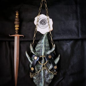 May include: A dagger with a metallic handle and a pointed blade next to a textured green dragon-scale-like object suspended by gold chains from a decorative wall mount. The green object features black leather straps with gold rivets and a central ring.