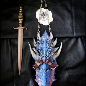 Puede incluir: Una daga con mango metálico y hoja puntiaguda junto a un escudo texturizado de escamas de dragón en azul y morado. El escudo presenta protuberancias afiladas, un anillo central, correas de cuero marrón con remaches y una cadena.