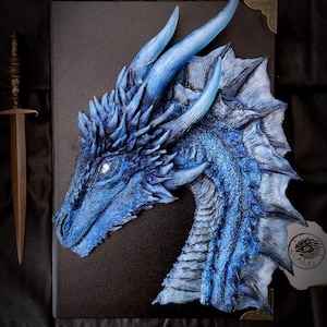 Peut inclure: Un livre rectangulaire noir orné d'une sculpture tridimensionnelle de tête de dragon bleu sur la couverture. Le dragon présente des écailles texturées, des cornes saillantes et un œil pâle. Une petite dague brune est posée à gauche, logo SCALES visible.