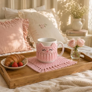 May include: A white ceramic mug featuring a light pink crocheted cozy with a cat face design and fringe trim, resting on a matching pink crocheted square coaster with fringe edges, accompanied by a croissant and two strawberries on a plate.